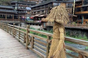 A traditional wooden bridge with a thatched figurehead, leading to a row of traditional buildings with multiple stories and a wooden fence. - Xijiang miao village listen to tao xuan hotel (Leishan)
