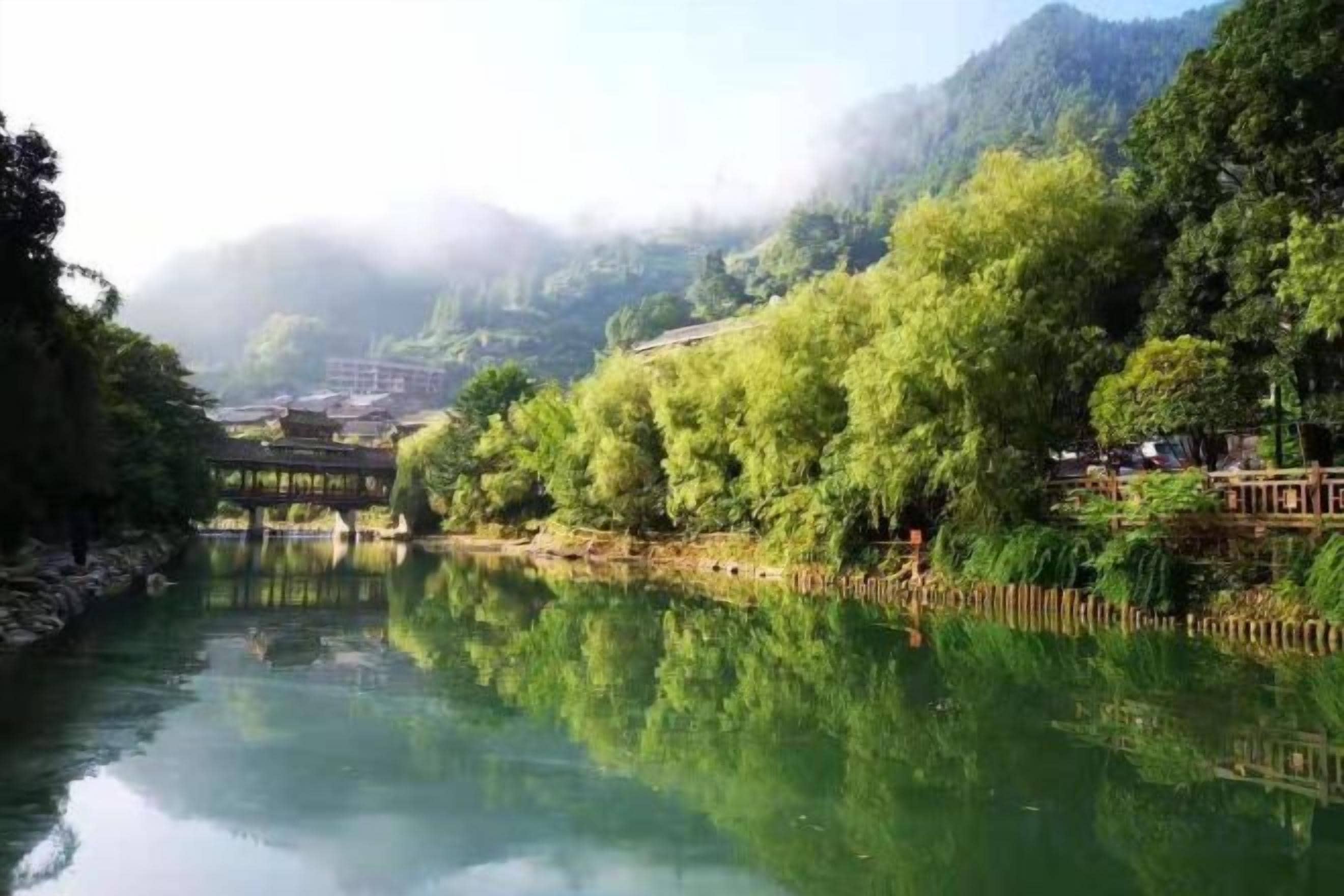 A river with a bridge, surrounded by lush greenery and a misty mountain backdrop.