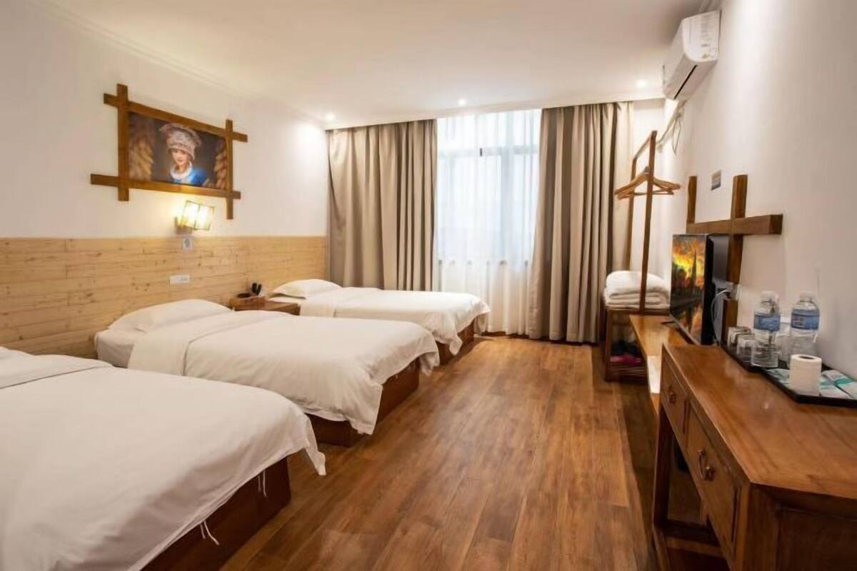 Ein Hotelzimmer mit drei Betten, Holzboden, einem Holzkopfteil, einem Gemälde, einem Spiegel, einer Holzkommode und einem Fenster mit Vorhängen.