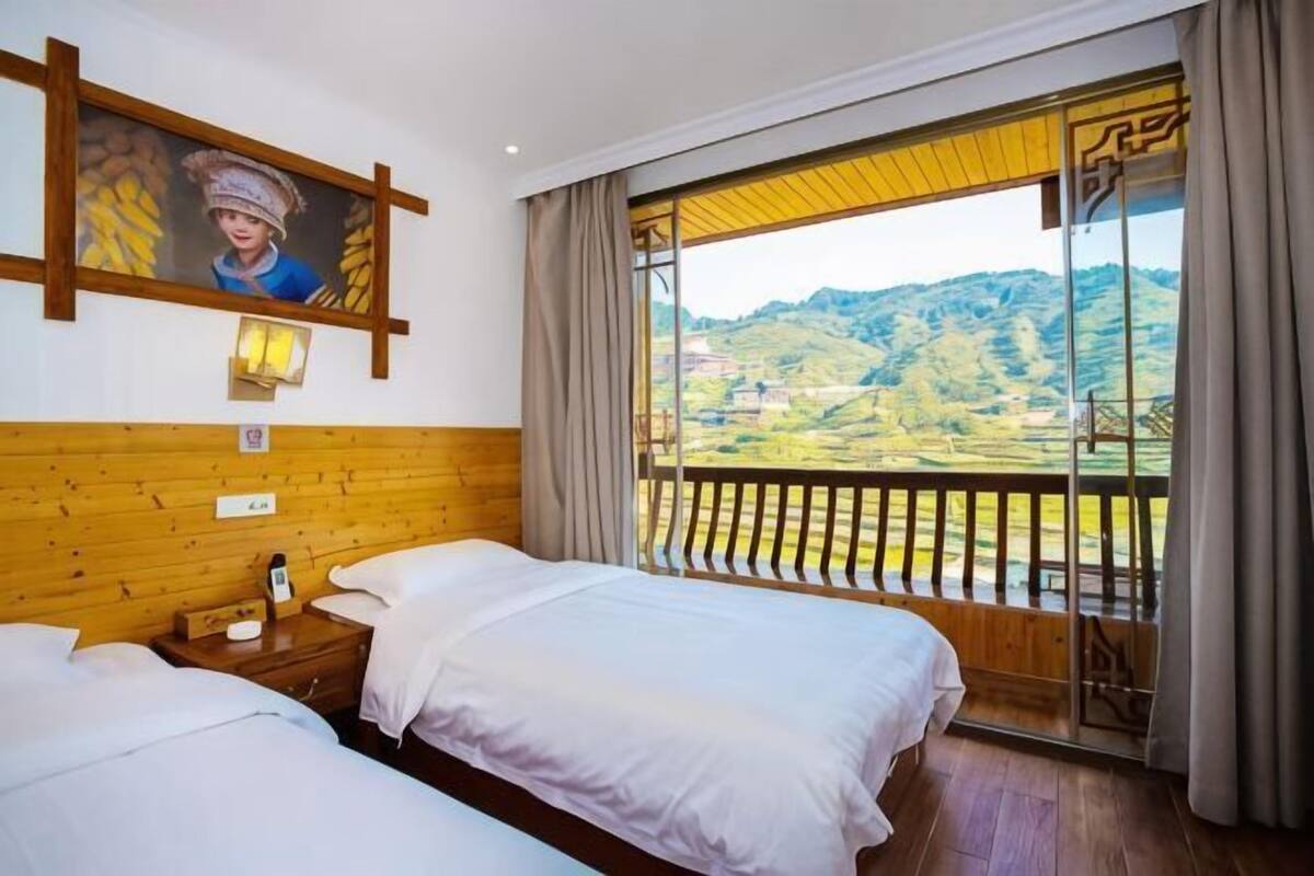 Ein Hotelzimmer mit zwei Betten, einem Holzkopfende, einem Gemälde, einem Nachttisch und einem großen Fenster mit Blick auf eine Berglandschaft.