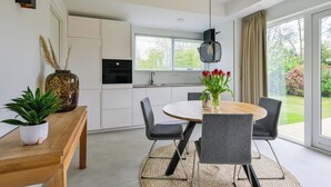 A modern kitchen with white cabinets, a dining area with a round table and grey chairs, and a large window with a view of greenery. - Eureka 36 Bungalow for 4-guests With Garden (Sint Maartensvlotbrug)