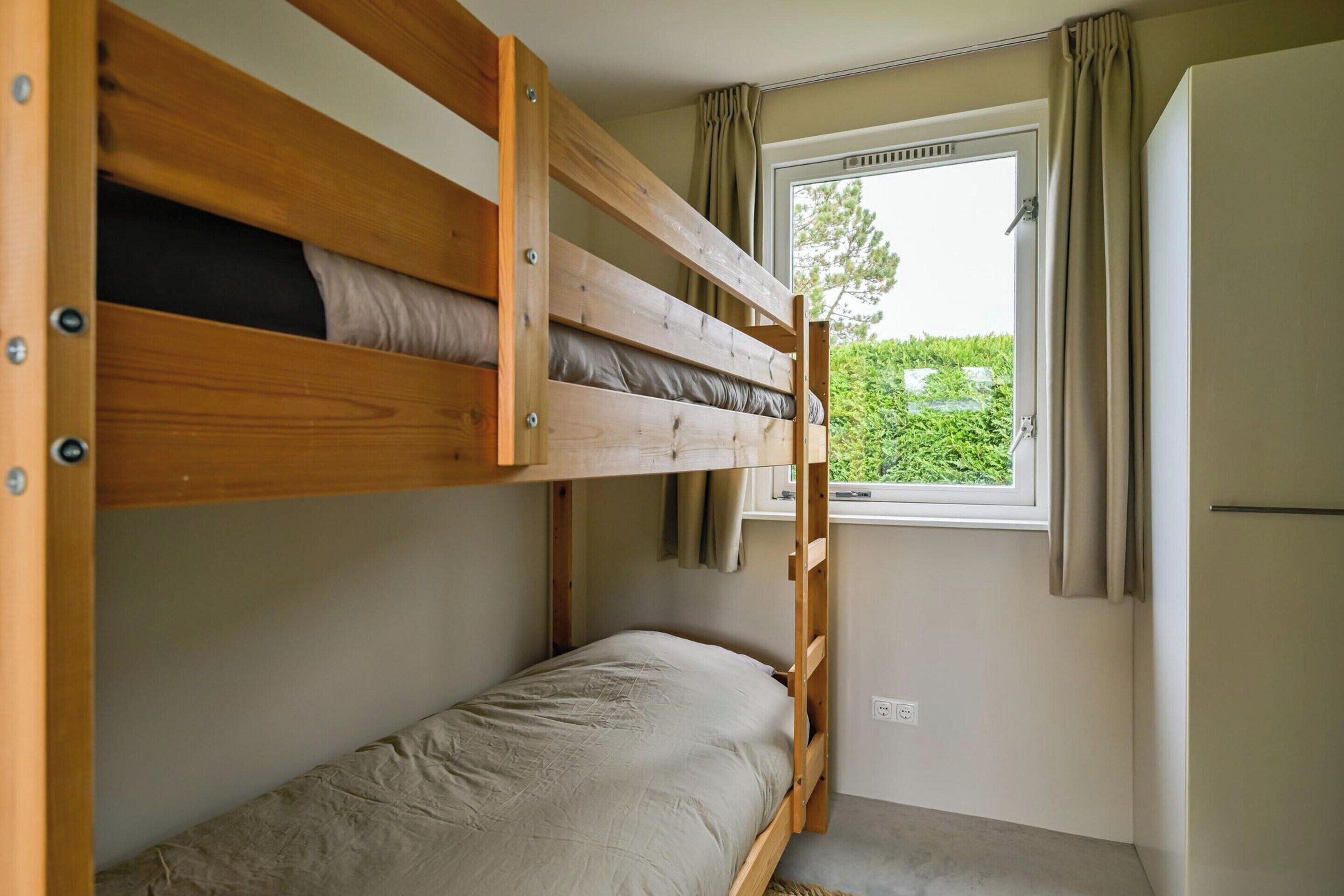 A bunk bed room with a window and curtains, a small kitchenette, and a view of greenery outside.