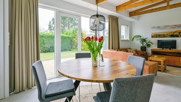 A modern living room with a round wooden dining table, grey upholstered chairs, a sofa, and a television on a wooden stand. - Eureka 36 Bungalow for 4-guests With Garden (Sint Maartensvlotbrug)