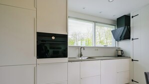 A modern kitchen with white cabinetry, a built-in oven, a sink, and a window with blinds. - Eureka 36 Bungalow for 4-guests With Garden (Sint Maartensvlotbrug)