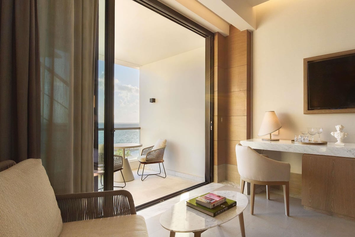 A modern hotel room with a balcony, a dining area, and a seating area with a book on the table.
