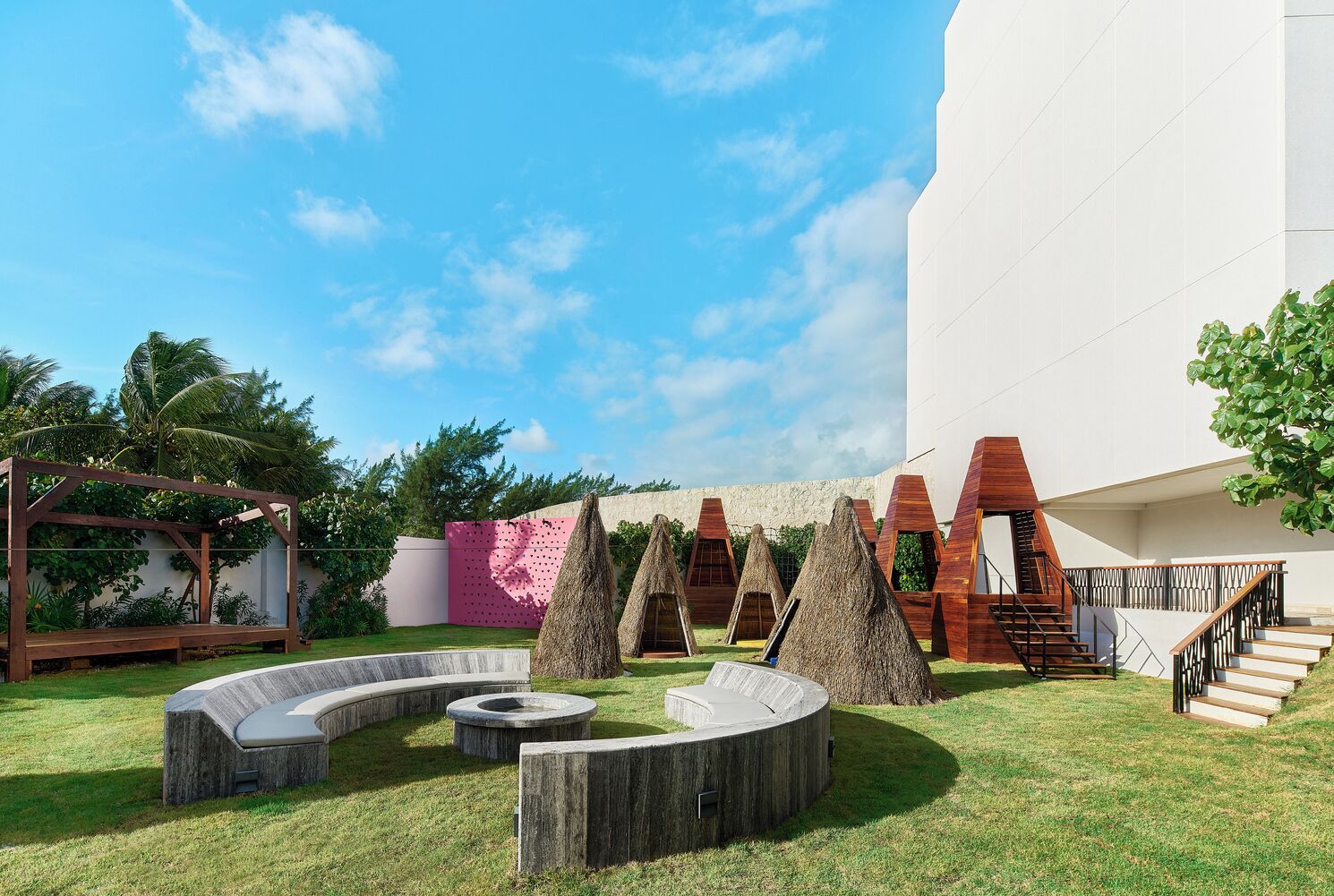 Un espacio exterior moderno con una pérgola de madera, una pared rosa con diseño de cuadrícula y varias estructuras cónicas.