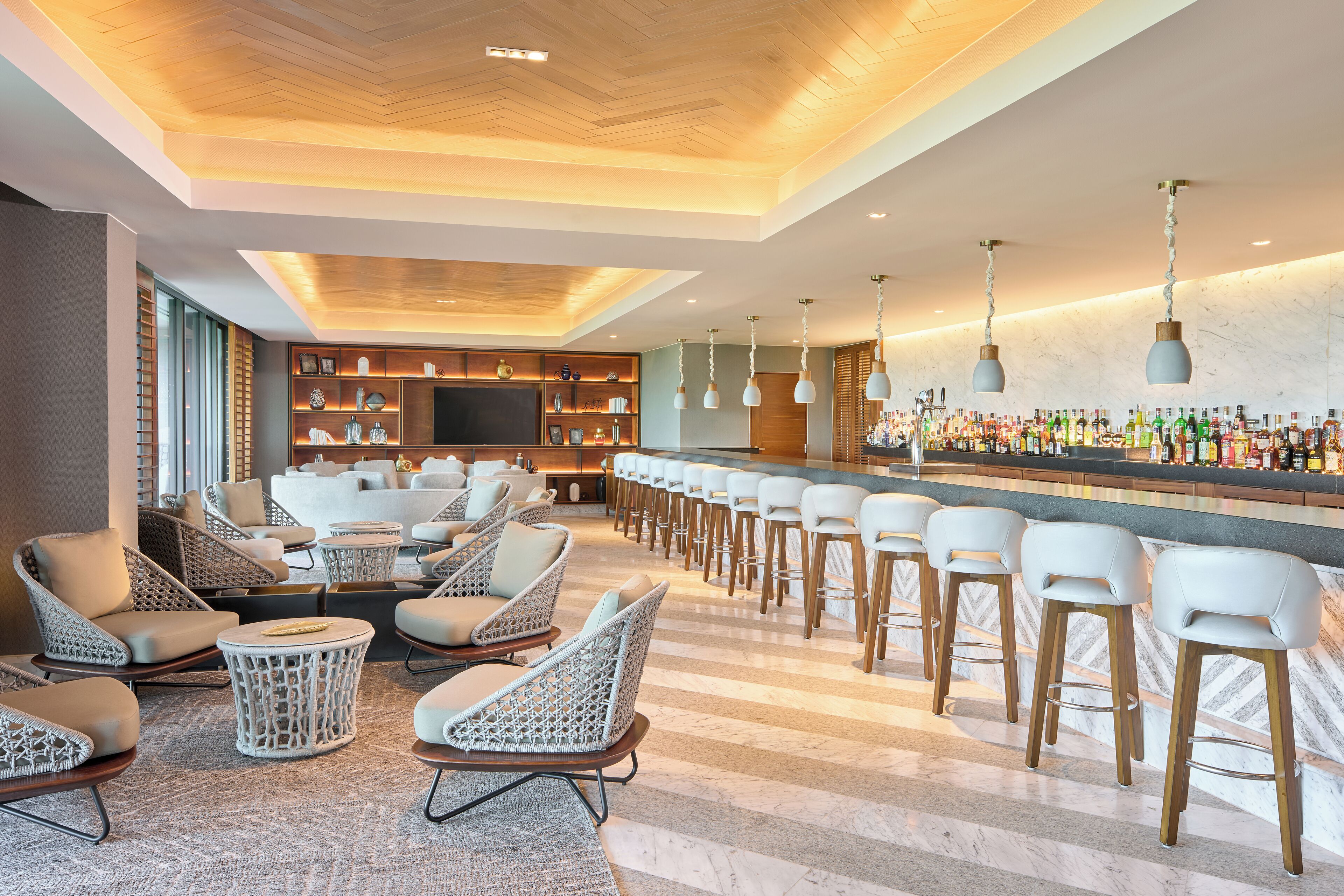 Una zona de bar con sillas y taburetes de mimbre, una barra alargada con botellas, y un techo con paneles de madera y luces colgantes.