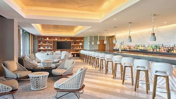 Una zona de bar con sillas y taburetes de mimbre, una barra alargada con botellas, y un techo con paneles de madera y luces colgantes.
