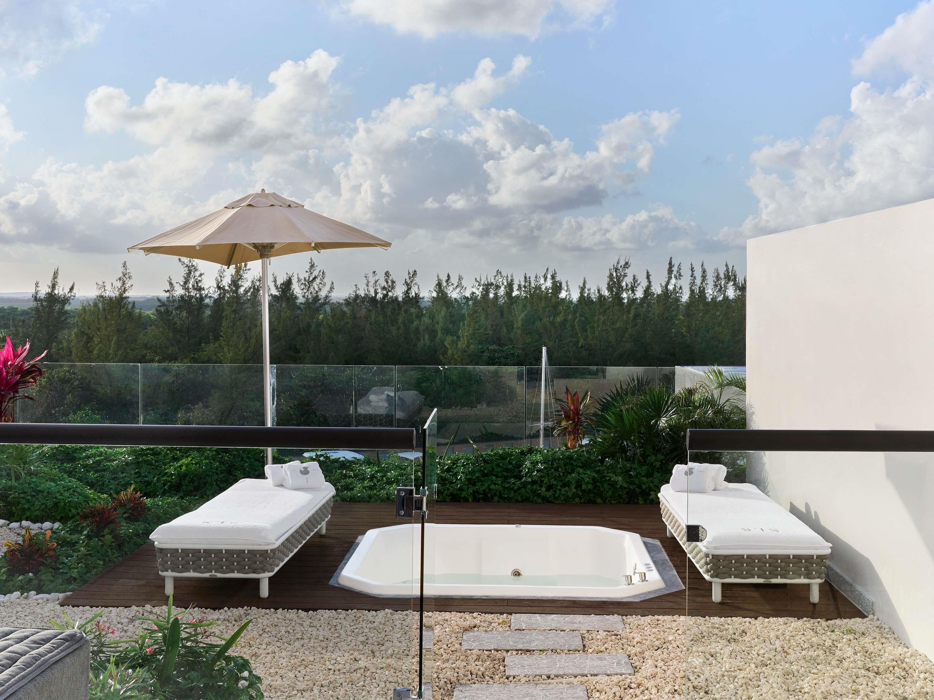 A rooftop terrace with two lounge chairs, a hot tub, and a glass railing overlooking a forested area.