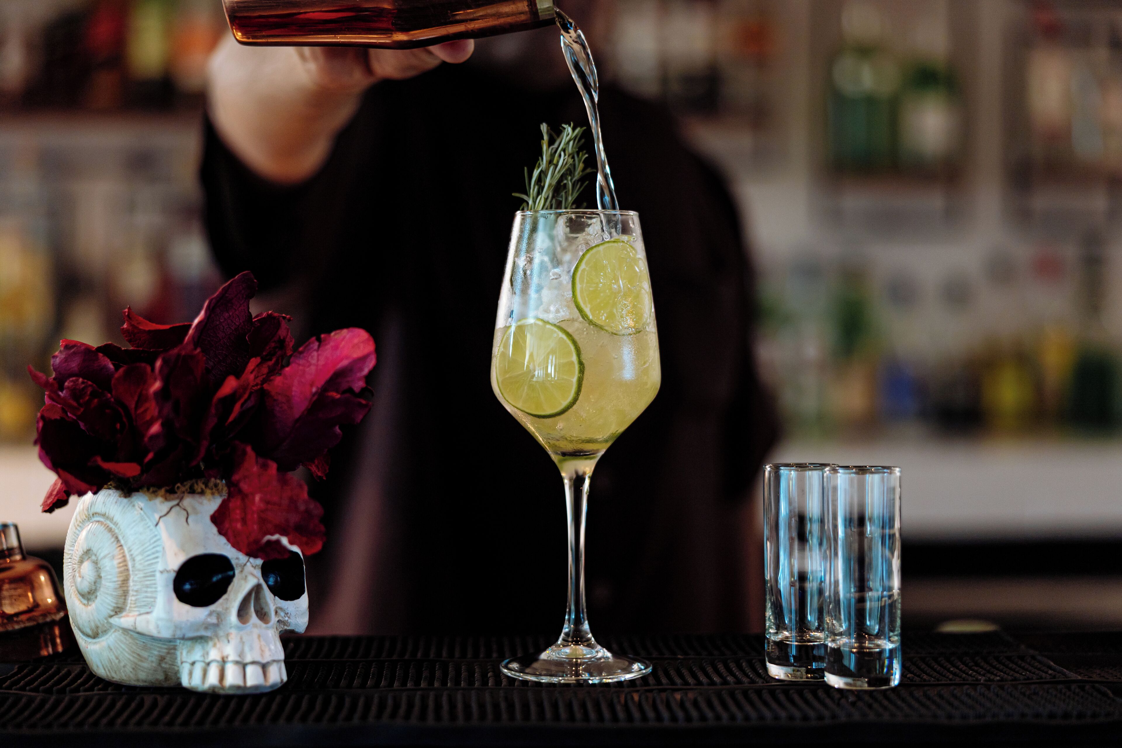 A bar setting with a skull-shaped vase containing red flowers, a cocktail glass with lime and rosemary, and two empty glasses on a bar mat.
