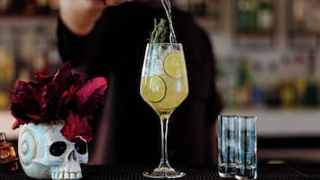A bar setting with a skull-shaped vase containing red flowers, a cocktail glass with lime and rosemary, and two empty glasses on a bar mat.