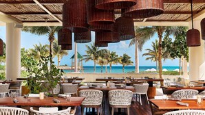An outdoor dining area with wicker furniture and hanging wicker lamps, overlooking a beach with palm trees and the ocean. - SLS Playa Mujeres – ALL Inclusive Collection (Punta Sam)