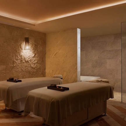 A spa room with two beds, a massage table, a bench, and a stone wall.