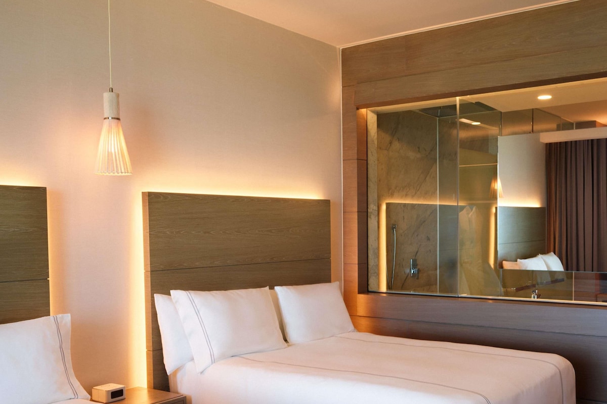 Habitación de hotel con dos camas, una mesita de noche, una lámpara, un espejo grande y zona de ducha.