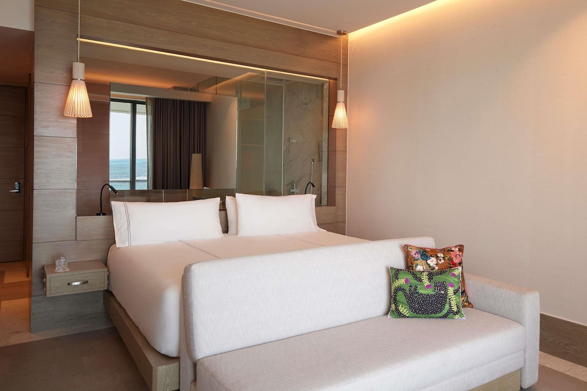 Una habitación de hotel moderna con una cama grande, un sofá, una mesita de noche, un espejo y vistas al mar.