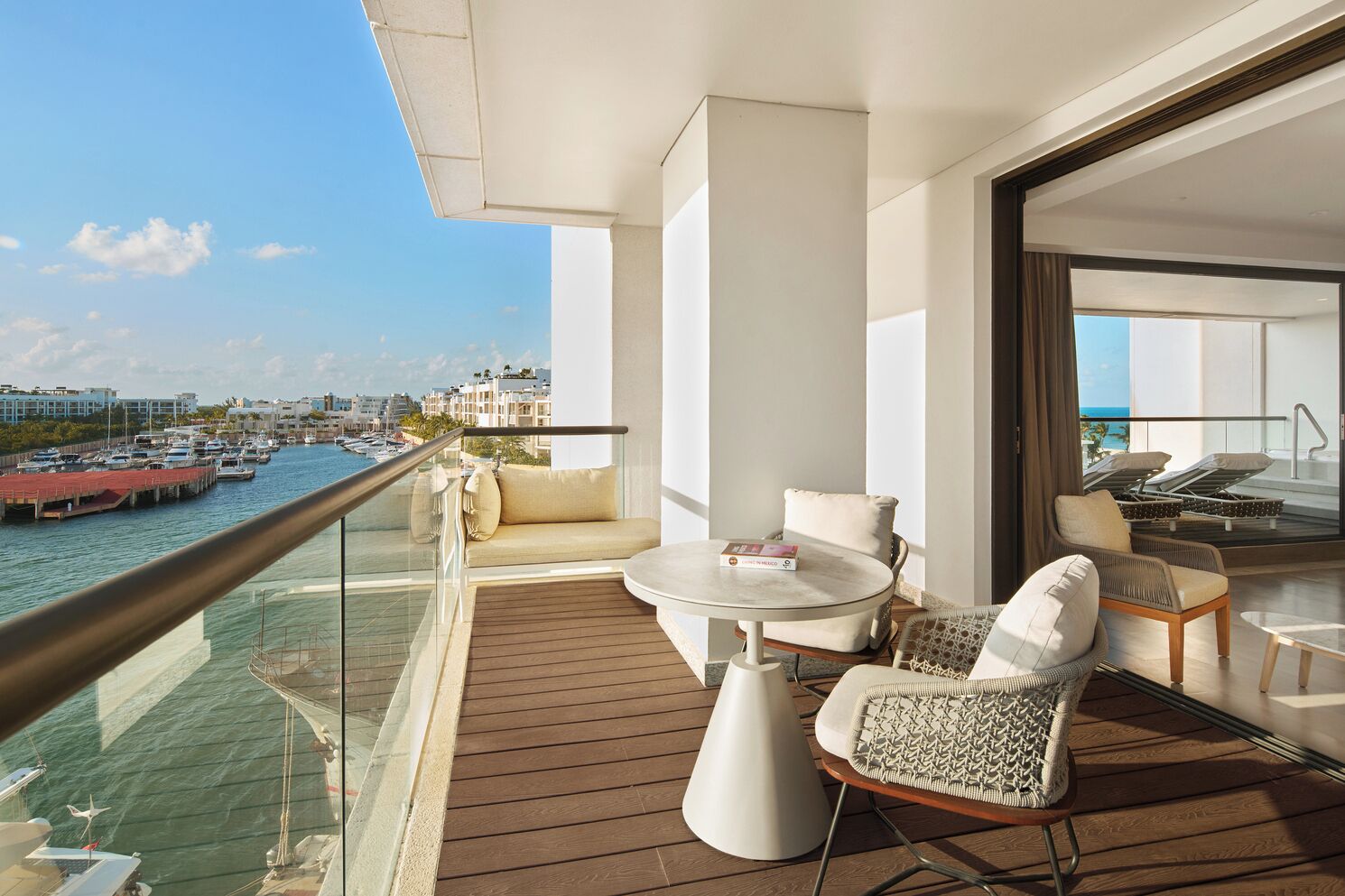 A balcony with a wooden deck, a round table, and two chairs. A glass railing separates the balcony from the water.