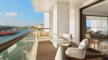 A balcony with a wooden deck, a round table, and two chairs. A glass railing separates the balcony from the water.