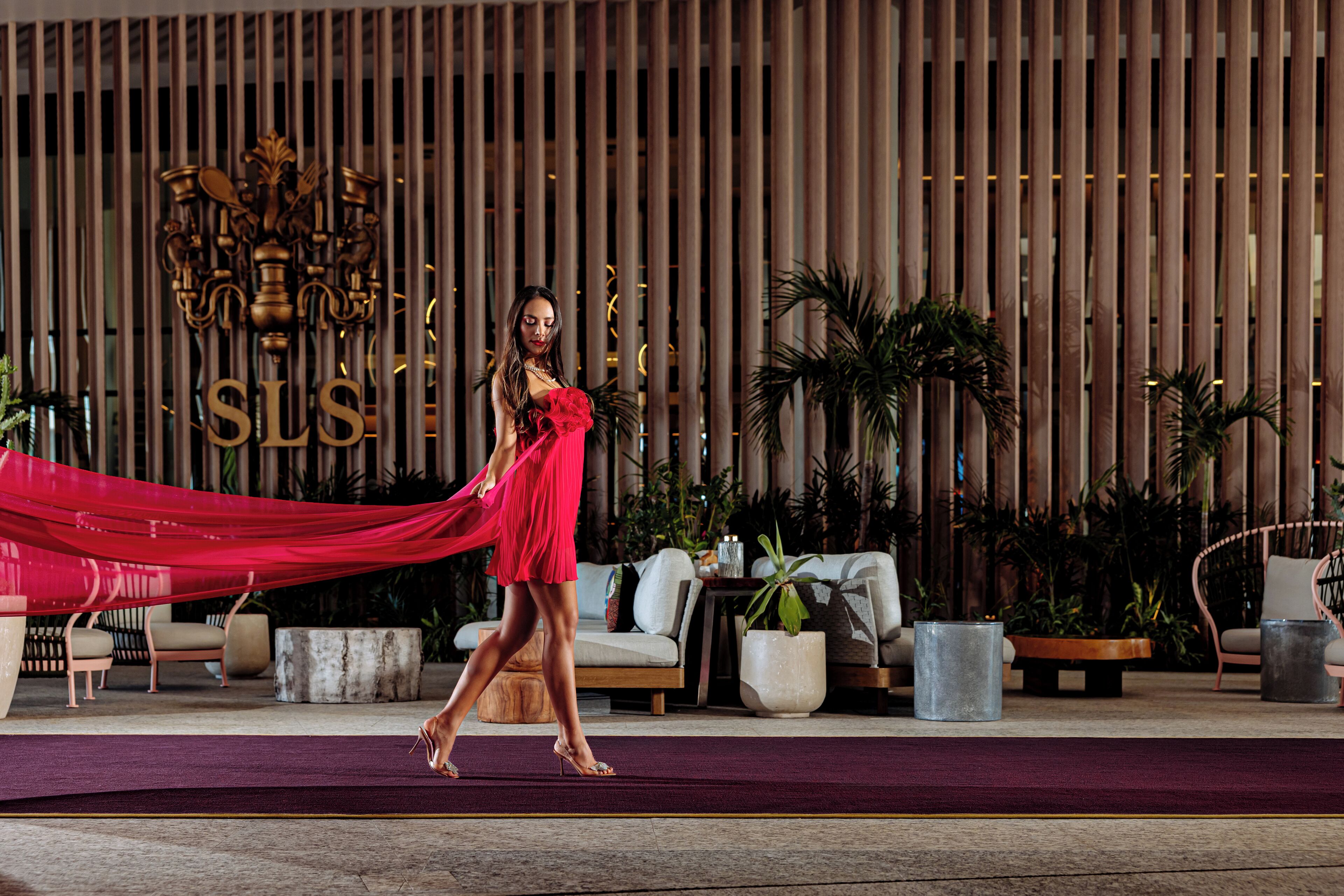 Una mujer con un vestido rojo caminando sobre un escenario con un fondo que incluye un panel de madera vertical y las letras 'SLS'.