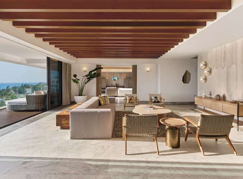 Una sala de estar moderna con un sofá grande, dos sillones, una mesa de centro y una mesa de comedor. Se aprecia vista al océano a través de grandes ventanales.
