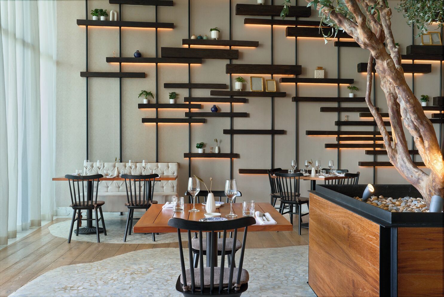 A modern restaurant interior with wooden tables and chairs, a large tree planter, and shelves with decorative items.