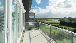 A balcony with a glass railing overlooking a grassy area and a road. - Strandleven 106 Apartment in Groote Keeten (Callantsoog)