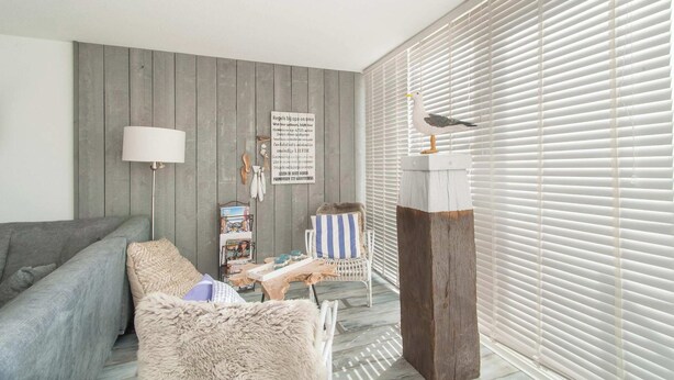 A living room with a grey sofa, a white armchair, a wooden wall, and a window with blinds. - Strandleven 106 Apartment in Groote Keeten (Callantsoog)
