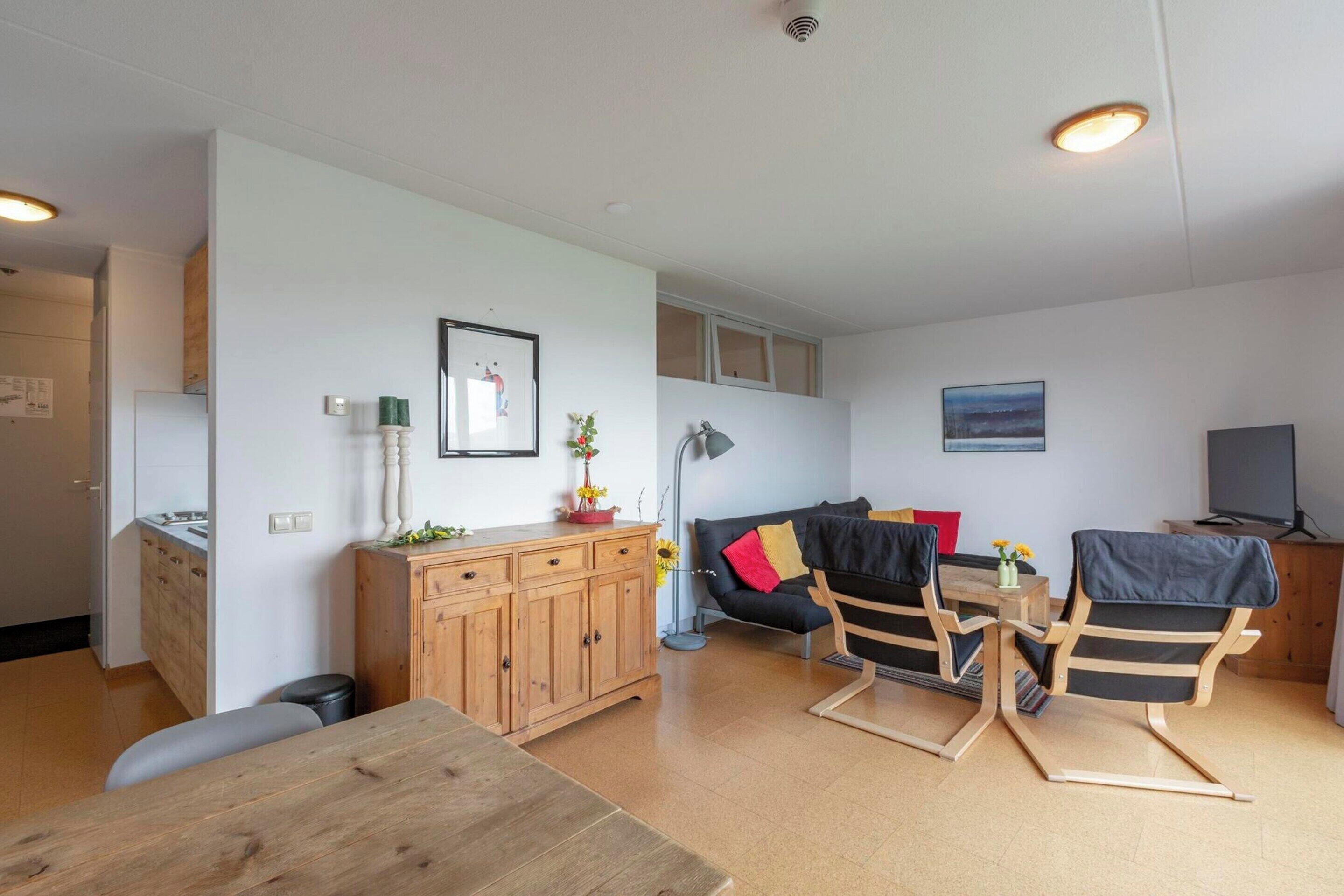 Una sala moderna con una mesa de comedor de madera, un sofá y dos sillones. Hay una zona de cocina con armarios y un refrigerador.