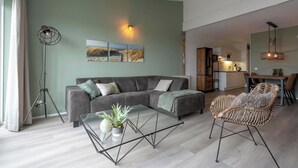 A modern living room with a sofa, a glass coffee table with a plant, a wicker chair, and a dining area in the background. - Strandslag 203 With Dunes View Balcony (Julianadorp)