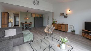 A modern living room with a grey sofa, a glass coffee table, a wicker chair, a dining area with a table and chairs, a kitchen in the background, and a TV stand with a flat-screen TV. - Strandslag 203 With Dunes View Balcony (Julianadorp)