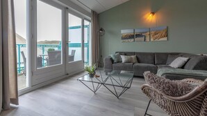 A spacious living room with a large window, a grey sofa, a glass coffee table, and a wicker chair. - Strandslag 203 With Dunes View Balcony (Julianadorp)