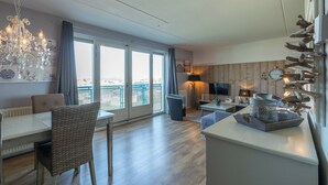 A modern living room with a dining area, large windows, a chandelier, and a television. - Strandslag 193 Charming Apartment for 4-person (Julianadorp)