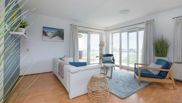 A modern living room with a white sofa, a wooden chair, and a blue armchair. There is a large window with curtains, a small table with a lamp, and a wicker stool. - Residence Juliana 40 With Dune Views (Julianadorp)