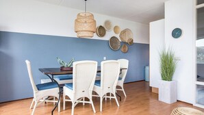 A dining area with a wooden table, white wicker chairs, a hanging light fixture, and a wall with decorative plates. - Residence Juliana 40 With Dune Views (Julianadorp)