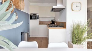 A compact kitchen with white cabinets, a stove, and a sink. There are two white chairs and a trash can. - Residence Juliana 40 With Dune Views (Julianadorp)