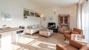 Una sala de estar moderna con un sofá, una mesa de centro y un televisor. Hay una zona de cocina con mostrador y sillas.