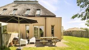 A modern wooden house with a thatched roof, a patio with a table and chairs, and a wooden fence in the background. - Vossehoeck 3 Apartment With Nature Reserve View (Callantsoog)