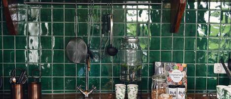 A kitchen with a green tiled backsplash, hanging utensils, a stainless steel sink, and various kitchen items on the counter.