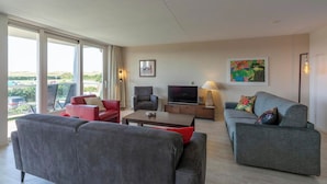 A modern living room with grey sofas, a red armchair, a flat-screen TV, and a large window offering a view of the city. - Apartment in Groote Keeten Near Sandy Beach (Callantsoog)