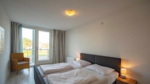 A hotel room with two beds, a chair, a nightstand with a lamp, and a view of the outdoors through a sliding door. - Apartment in Groote Keeten Near Sandy Beach (Callantsoog)