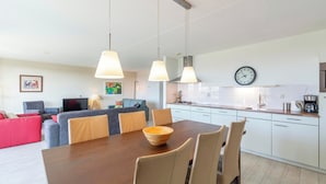 A modern living room with a dining table, chairs, and a kitchen area with white cabinets and a clock on the wall. - Apartment in Groote Keeten Near Sandy Beach (Callantsoog)