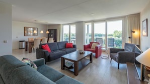 A spacious living area with a dark-colored sofa, a red armchair, and a grey sofa. There is a wooden coffee table in the center, and a lamp on a side table. Large windows provide a view of the outdoors. - Apartment in Groote Keeten Near Sandy Beach (Callantsoog)