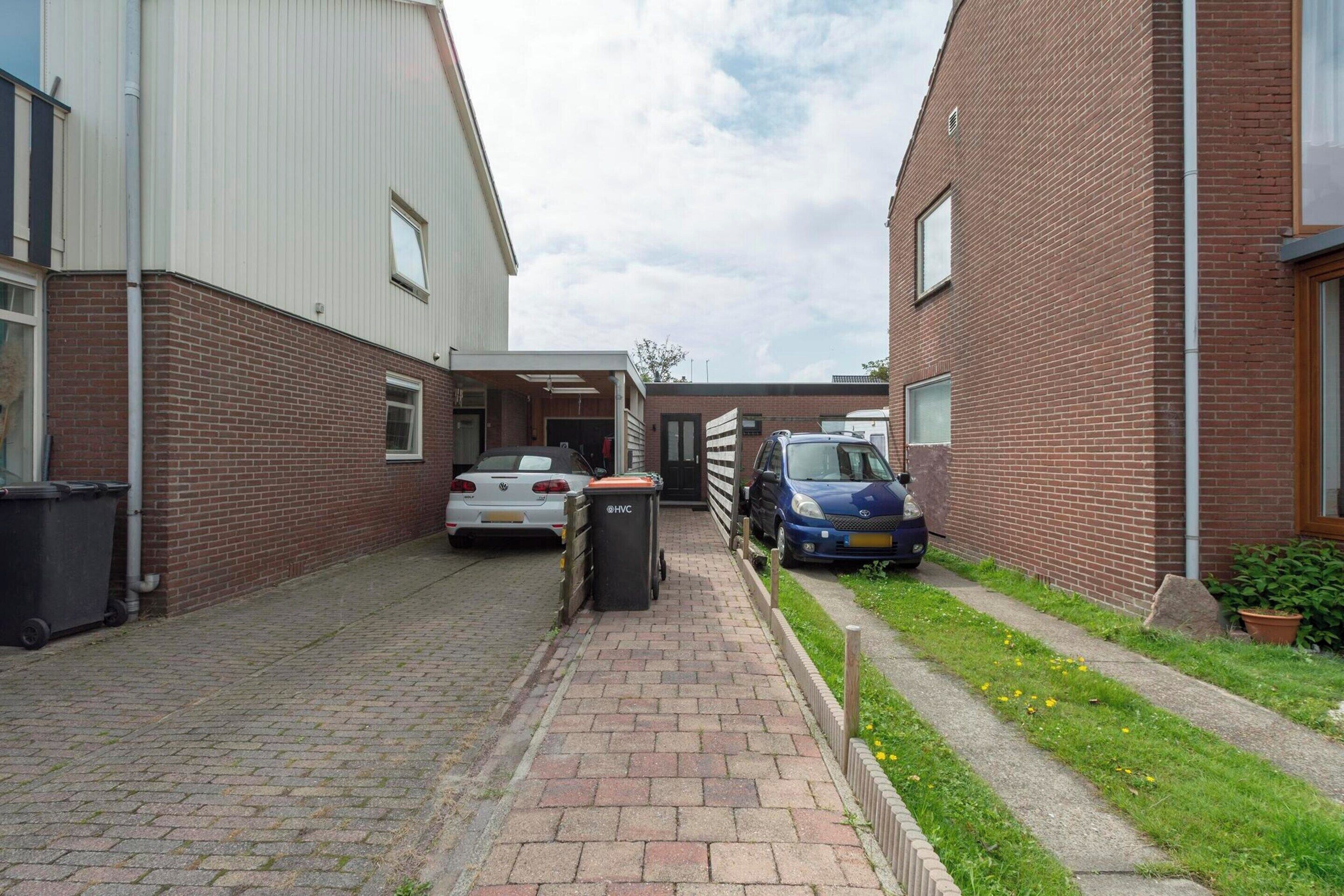 A narrow brick alleyway with two parked cars, a trash bin, and a pathway leading to a building with a white exterior.
