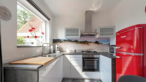 A modern kitchen with white cabinets, a red refrigerator, a built-in oven, and a window with a view of a beach. - Stuijvezandeweg 7Z for 2 Guests Near Centre (Callantsoog)