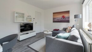 A modern living room with a flat-screen TV, grey sofa, and a painting of a sunset. - Stuijvezandeweg 7Z for 2 Guests Near Centre (Callantsoog)
