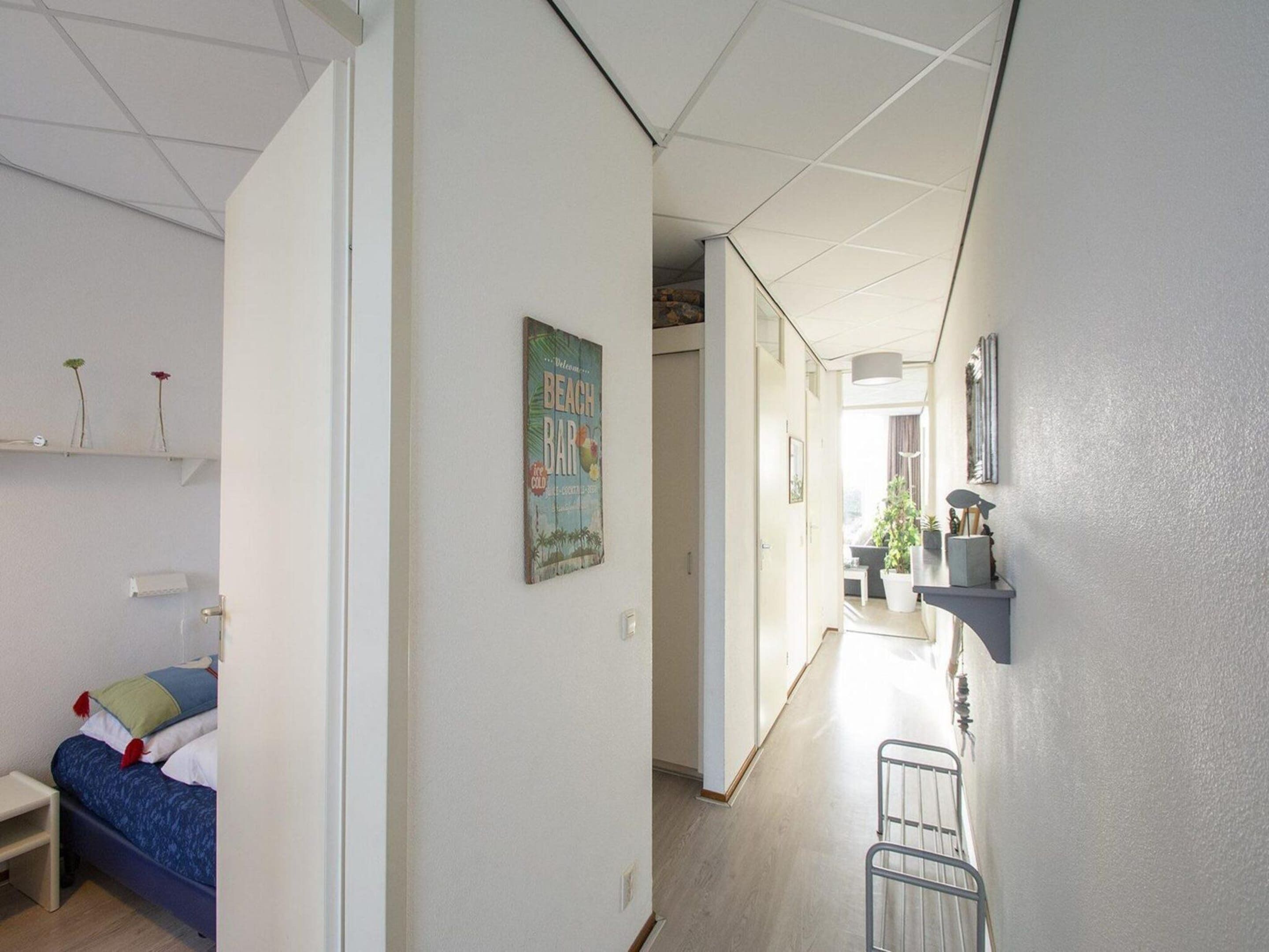 A hallway with a bed, a wall-mounted shelf with flowers, a poster, and a small table.