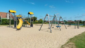 A playground with a yellow slide and swings, set on sandy ground with houses in the background. - Apartment 189 in Julianadorp aan Zee (Julianadorp)