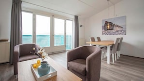 A modern room with a dining table, two armchairs, a coffee table with a vase and candles, and a large window with a view of the sea. - Apartment by the Dunes Near the Sea (Julianadorp)