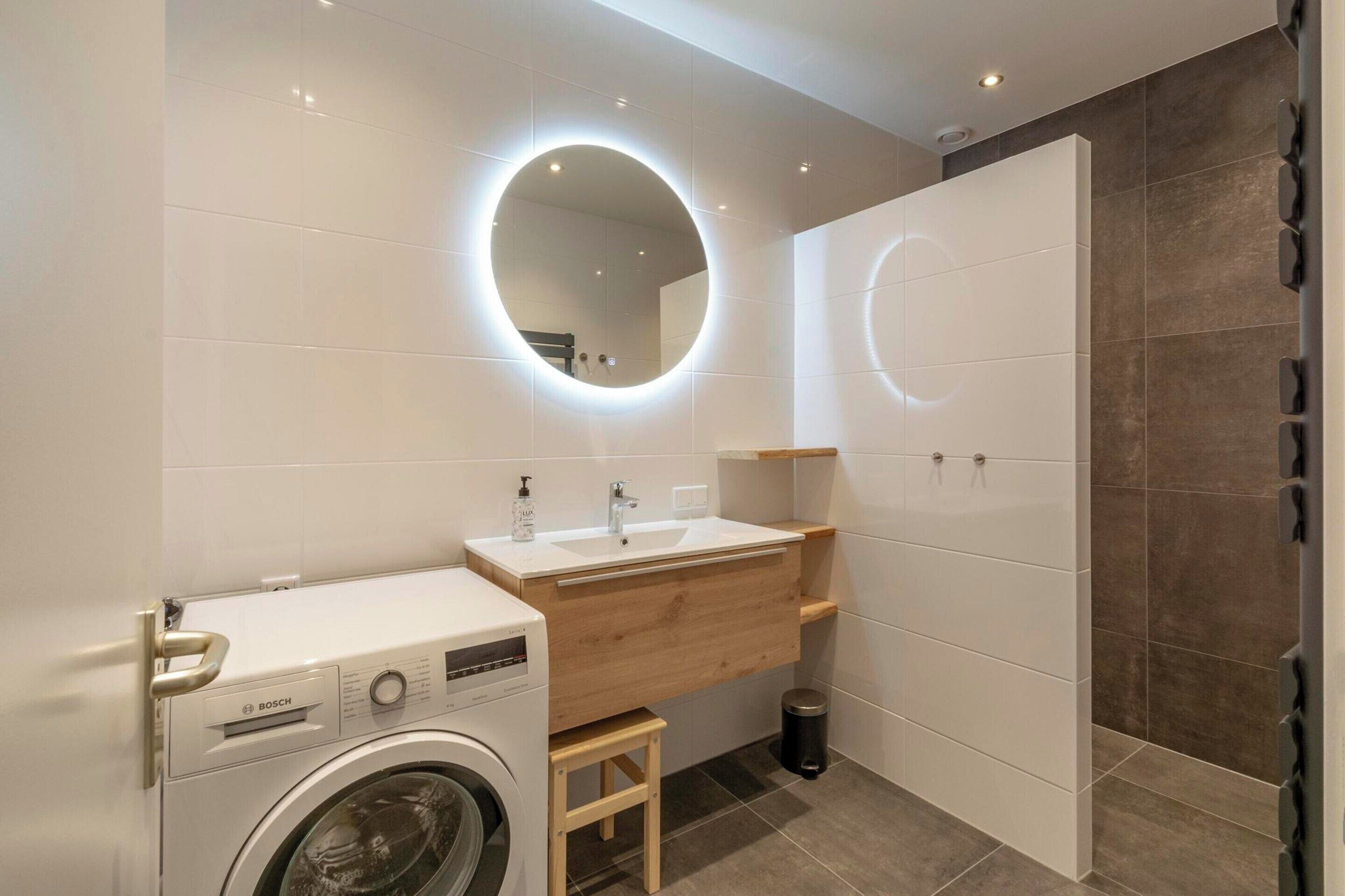 Un baño moderno con una lavadora Bosch, un tocador de madera, un espejo redondo y un perchero para toallas.