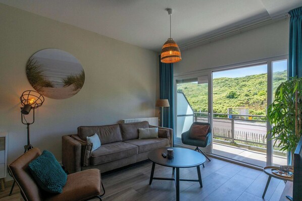 A modern living room with a sofa, armchair, and round coffee table. A large window offers a view of greenery outside.
