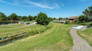 A park with a paved path, grassy areas, a small water channel, and a playground. - Apartment in Julianadorp by the Sea (Julianadorp)